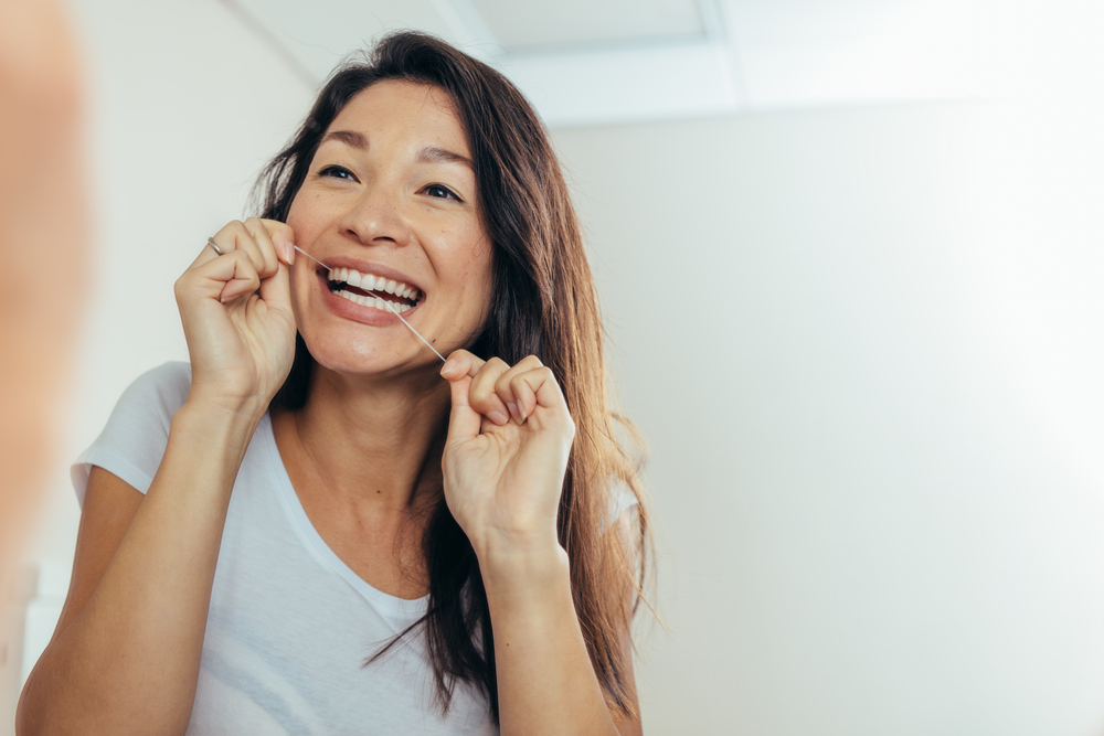 Woman smiling while flossing teeth in a bathroom, emphasizing daily oral hygiene and preventive dental care.